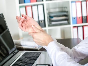kobieca dłoń masuje nadgarstek drugiej dłoni, w tle laptop i biuro